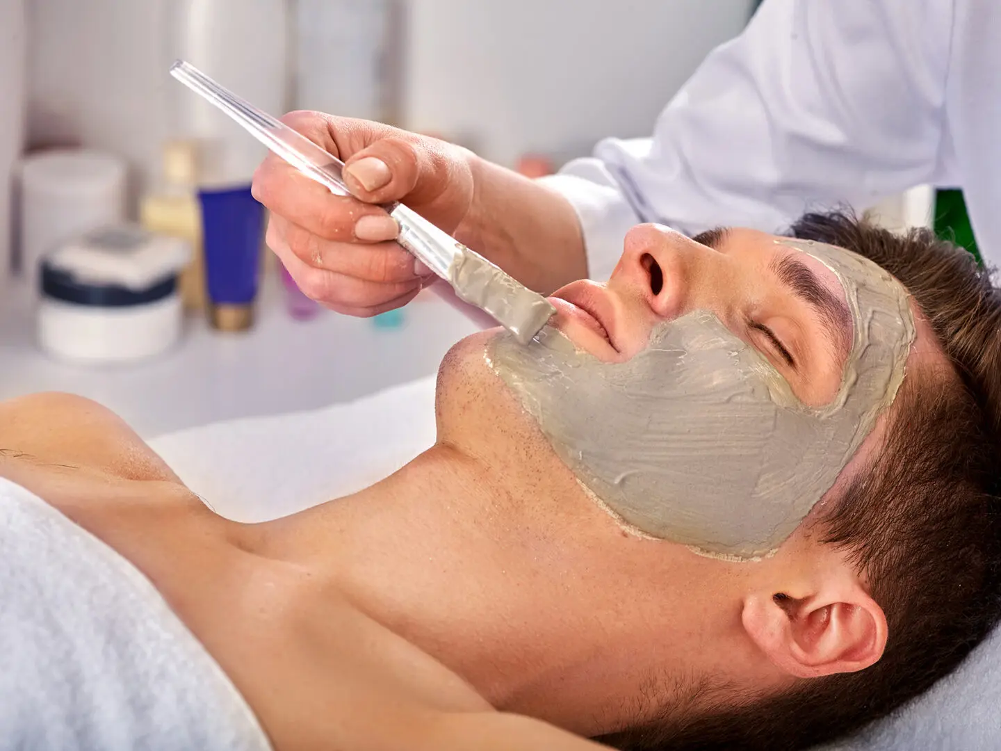 Mud facial mask of man in spa salon. Massage with clay full face in therapy room.